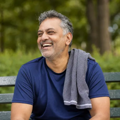 A man in his fifties laughing on a park bench in workout clothes, a towel over his shoulder